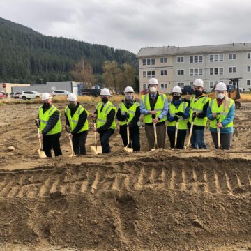 Eight adults wearing white hard hats and yellow safety vests hold golden shovels on the site of the future Teal Street Center