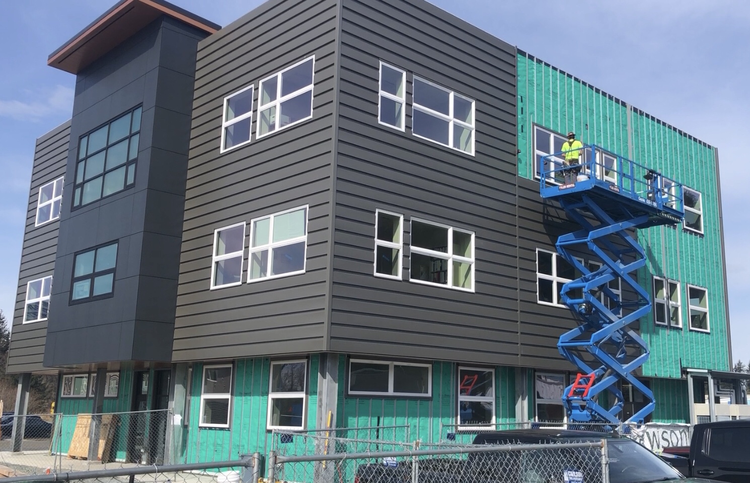 Teal Street Center in progress. There is grey siding on most of the 3-story building, plus some green board. There's a blue lift with a person standing on it working on the third floor
